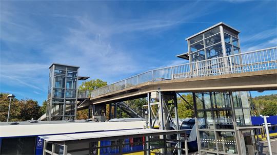 Zwei gläserne Aufzüge mit einer darüber verlaufenden Fußgängerbrücke vor blauem Himmel