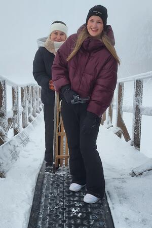 Zwei Personen in Winterkleidung stehen auf einem schneebedeckten Laufband, eine hält einen Schlitten.