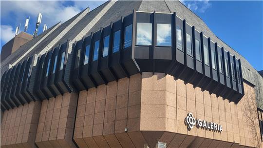 Eckansicht eines mehrstöckigen Gebäudes mit brauner Fassade und schwarzen, vorstehenden Fenstern unter blauem Himmel