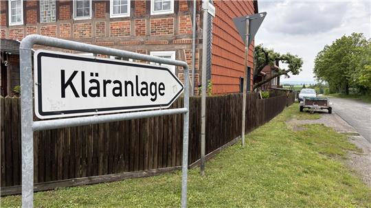 Wegweiser mit der Aufschrift 'Kläranlage' vor einem Zaun und einem Backsteinhaus an einer Landstraße mit parkendem Auto und Bäumen im Hintergrund
