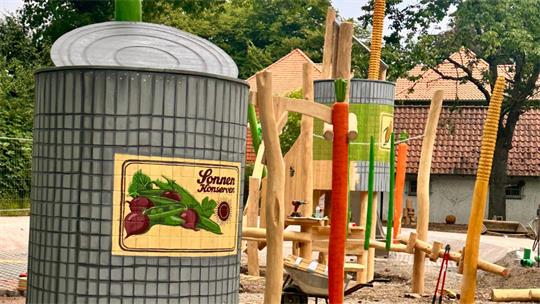 Mülleimer mit Holzschild und Spielplatz mit Klettergerüst im Hintergrund