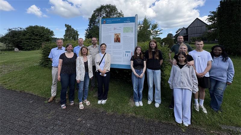 Gruppe von Menschen steht auf einer Wiese auf einem Friedhof vor einem großen Informationsschild bei leicht bewölktem Himmel