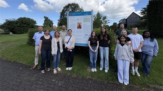 Gruppe von Menschen steht auf einer Wiese auf einem Friedhof vor einem großen Informationsschild bei leicht bewölktem Himmel