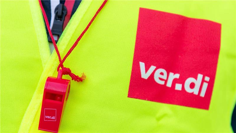 Eine gelbe Warnweste mit einem roten Quadrat drauf, auf dem „ver.di“ steht. Daneben hängt eine rote Trillerfeife.