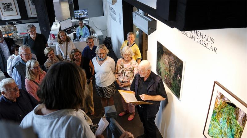 Gruppe von Menschen in einem Museum, die einem älteren Mann zuhören, der ein Buch oder eine Mappe zeigt.