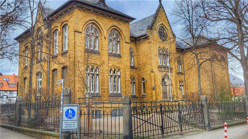 Historisches gelbes Backsteingebäude mit spitzbogigen Fenstern hinter einem schwarzen Metallzaun