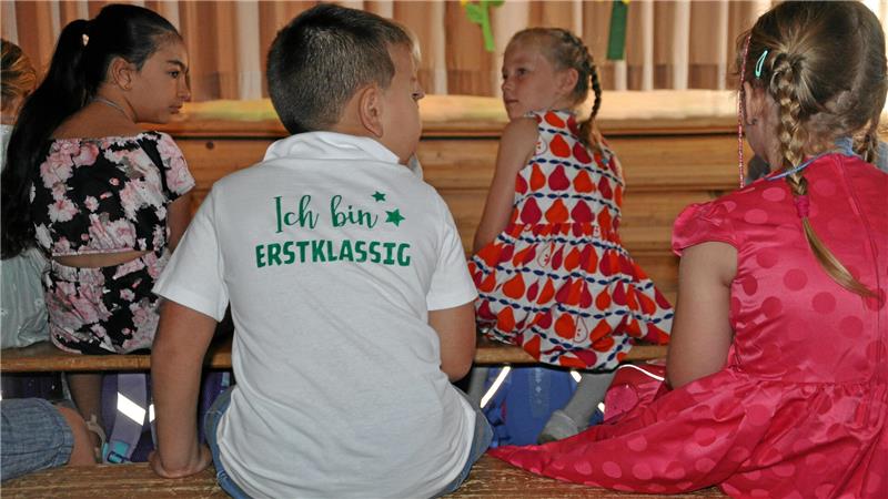 Vier Kinder sitzen auf einer Bank, eines trägt ein weißes T-Shirt mit der Aufschrift 'Ich bin ERSTKLASSIG'
