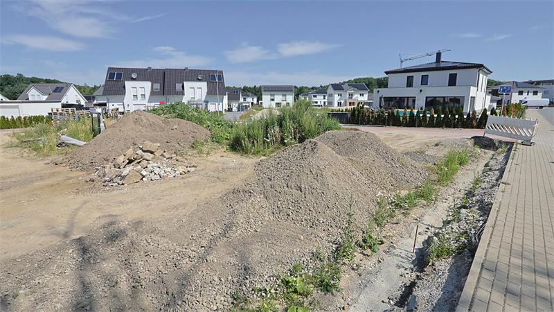 Baustelle mit mehreren Erdhaufen und Kieshaufen vor modernen Wohnhäusern unter blauem Himmel