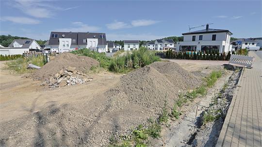 Baustelle mit mehreren Erdhaufen und Kieshaufen vor modernen Wohnhäusern unter blauem Himmel