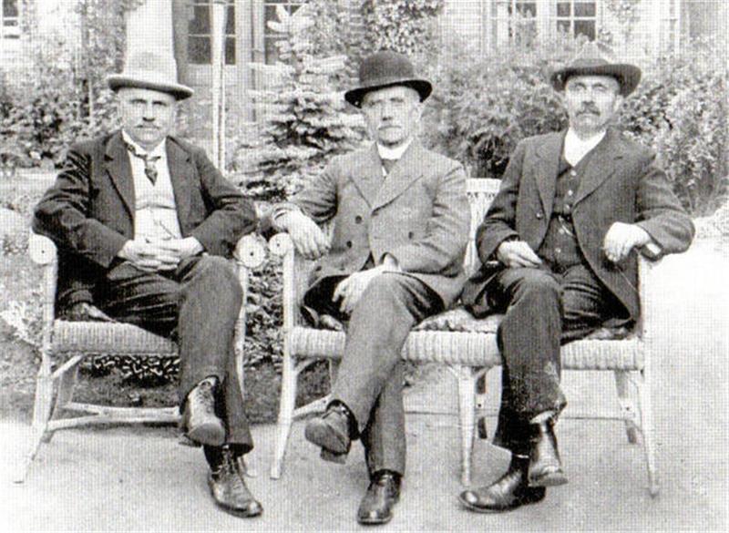 Tod in Odessa: Sohn eines Goslarer Zwangsarbeiterkindes stirbt Drei Männer in Anzügen und Hüten sitzen auf einem Gartenstuhl vor einem Gebäude mit Fenstern und Kletterpflanzen.