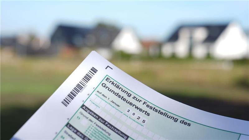 Das Symbolbild zeigt eine Grundsteuererklärung, im Hintergrund sind verschwommen Gebäude zu sehen.