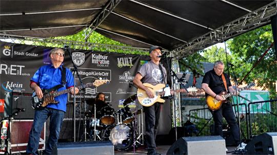 Vier Musiker spielen auf einer überdachten Bühne, zwei mit Gitarren, einer am Schlagzeug, einer mit Bass. Einer singt ins Mikrofon