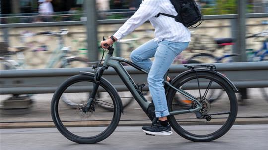 Person in weißem Hemd und Jeans fährt auf einem schwarzen Fahrrad auf einer Straße mit geparkten Fahrrädern im Hintergrund