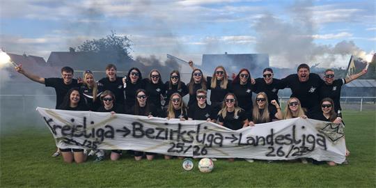 Gruppe von Fußballspielerinnen und -spielern auf einem Spielfeld mit einem Banner, auf dem 'Kreisliga -> Bezirksliga -> Landesliga' und Jahreszahlen stehen, einige halten Rauchfackeln in die Luft