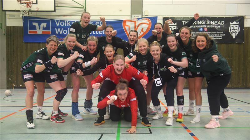 Gruppenfoto einer Handballmannschaft in Sportkleidung in einer Halle vor Werbebannern, einige Spieler in kämpferischer Pose, eine Person kniet vorne auf dem Boden.
