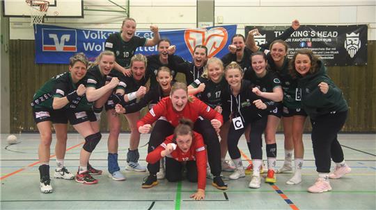 Gruppenfoto einer Handballmannschaft in Sportkleidung in einer Halle vor Werbebannern, einige Spieler in kämpferischer Pose, eine Person kniet vorne auf dem Boden.