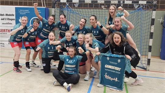 Gruppenfoto einer jubelnden Frauenhandballmannschaft in Sportkleidung in einer Halle vor einem Tor mit einem "Kempa"-Banner