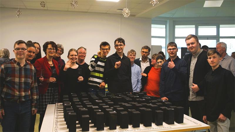 Gruppe von Menschen steht um einen Tisch mit vielen schwarzen rechteckigen Objekten in einem hellen Raum mit Fenstern und Schneeflockendekorationen an der Decke.