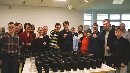 Gruppe von Menschen steht um einen Tisch mit vielen schwarzen rechteckigen Objekten in einem hellen Raum mit Fenstern und Schneeflockendekorationen an der Decke.