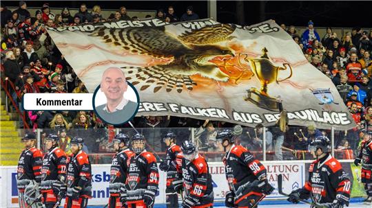 Eishockeyspieler in schwarz-roten Trikots auf dem Eis vor einer Tribüne mit Fans, die ein großes Banner mit einem Falken, einem Pokal und dem Text 'REGIO-MEISTER OHNE FRAGE, UNSERE FALKEN AUS BRAUNLAGE'. halten