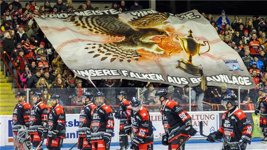 Eishockeyspieler in schwarzen und roten Trikots auf dem Eis vor einer großen Fan-Choreografie mit einem Falken, der einen Pokal hält, und dem Schriftzug 'Regio-Meister ohne Frage, unsere Falken aus Braunlage'