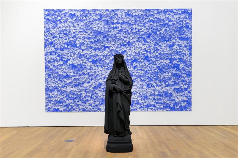 Die Heiligenfigur „St. Katharina“ aus Polyester und Acrylfarbe steht vor blauem Efeu (170 x 50 x 40 Zentimeter, Siebdruck, Kunststoff).