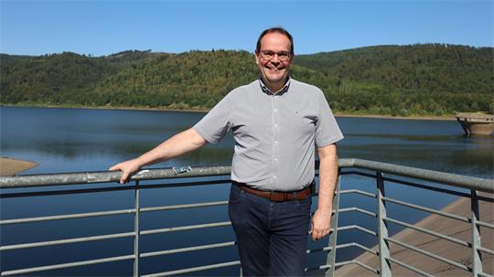 Mann mit Brille und gestreiftem Hemd lehnt an einem Geländer vor einem See und bewaldeten Hügeln im Hintergrund