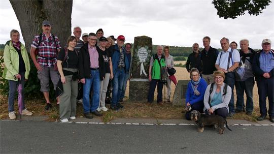 Gruppe von Menschen steht an einer Straße neben einem Baum und einem Stein mit einer Gedenktafel.