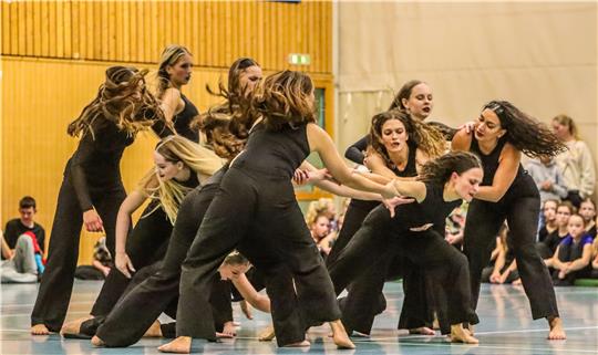 Gruppe von Tänzerinnen in schwarzer Kleidung führt eine synchronisierte Bewegung in einer Sporthalle vor Publikum aus.