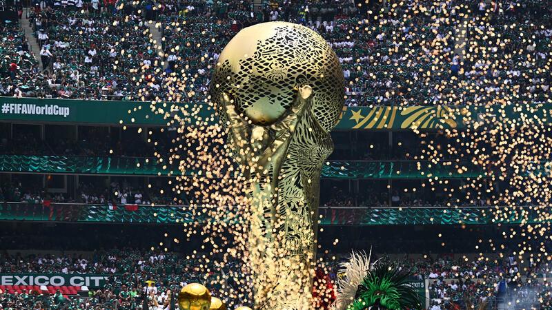 Goldene Skulptur einer menschlichen Figur, die eine große Kugel hält, vor einem Stadion mit Zuschauern und dem Schriftzug '#FIFAWorldCup'.