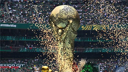 Goldene Skulptur einer menschlichen Figur, die eine große Kugel hält, vor einem Stadion mit Zuschauern und dem Schriftzug '#FIFAWorldCup'.