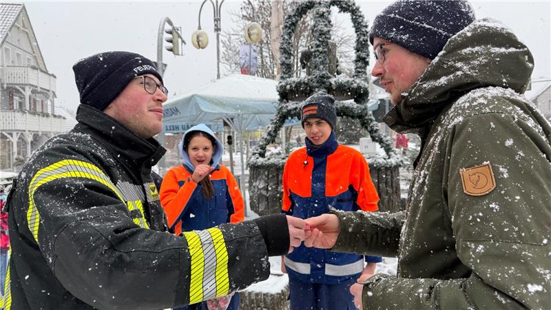 Vor den Augen von zwei Jugendfeuerwehrleuten verkauft ein Mann in Feuerwehrkleidung einem anderen ein Glücksschweinchen.