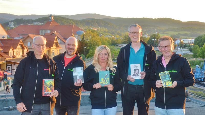 Fünf Personen stehen nebeneinander im Freien vor einer Landschaft mit Häusern und Hügeln und halten jeweils ein Buch in der Hand.