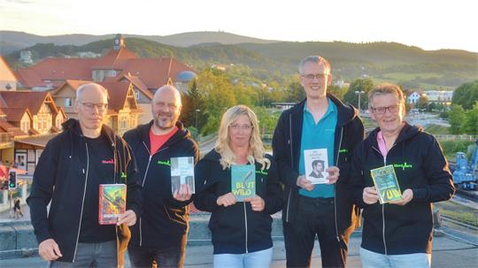 Fünf Personen stehen nebeneinander im Freien vor einer Landschaft mit Häusern und Hügeln und halten jeweils ein Buch in der Hand.