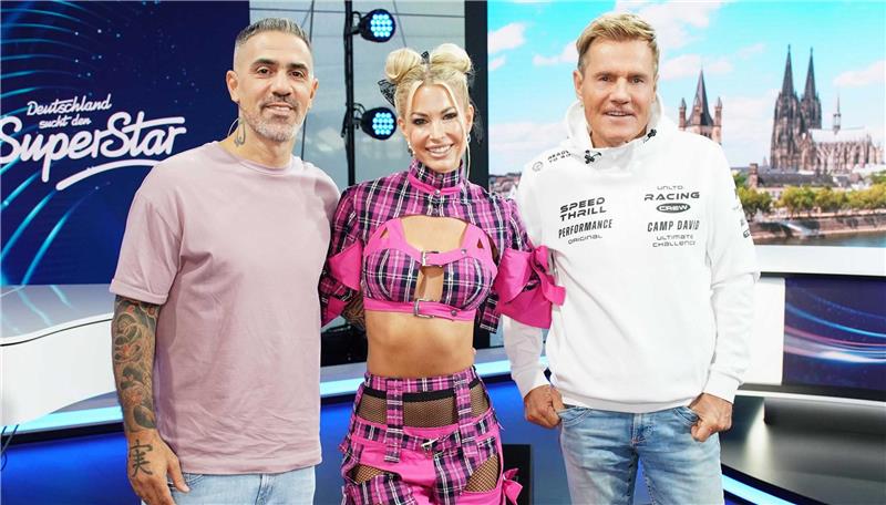 Ein Mann, eine Frau und ein weiterer Mann stehen in einem TV-Studio. Im Hintergrund steht "Deutschland sucht den Superstar" an der Wand und man sieht die Skyline der Stadt Köln.