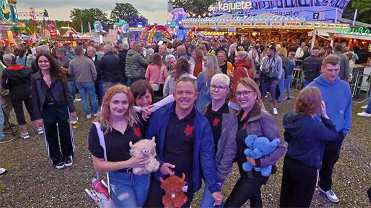 Gruppe von fünf Personen auf einem belebten Jahrmarkt, zwei halten Plüschtiere, im Hintergrund bunte Fahrgeschäfte und Menschenmengen