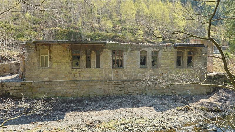 Verfallenes, langgestrecktes Steingebäude mit mehreren rechteckigen Fenstern in bewaldeter, hügeliger Landschaft