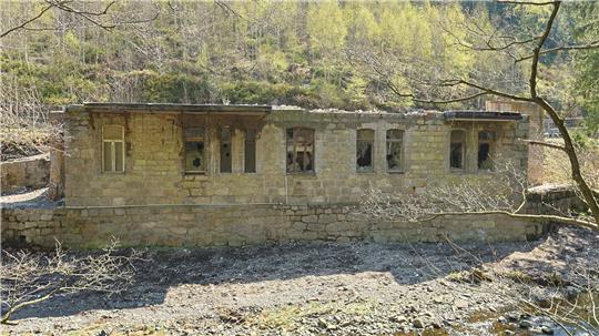 Verfallenes, langgestrecktes Steingebäude mit mehreren rechteckigen Fenstern in bewaldeter, hügeliger Landschaft