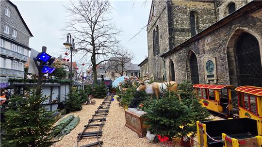 Weihnachtlich geschmückter Außenbereich mit Miniatureisenbahn, Tannenbäumen und einer lebensgroßen Rentierfigur neben einer alten Kirche.