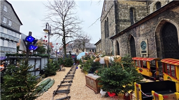 Weihnachtlich geschmückter Außenbereich mit Miniatureisenbahn, Tannenbäumen und einer lebensgroßen Rentierfigur neben einer alten Kirche.