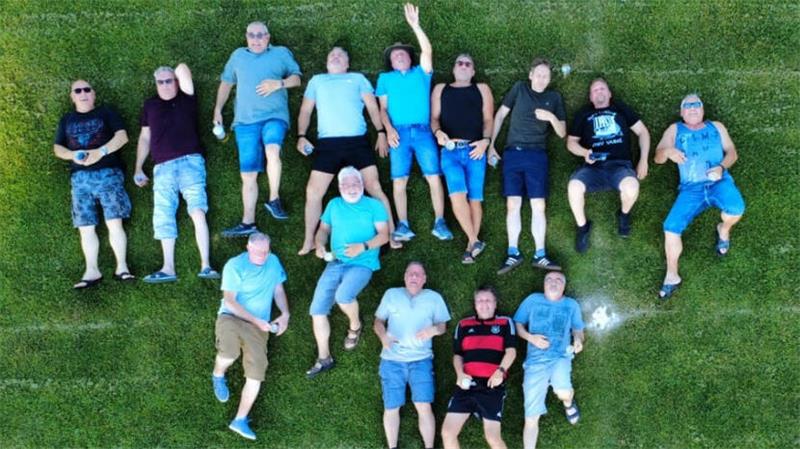 Gruppe von Männern liegt auf grünem Rasen, einige tragen T-Shirts und Shorts, einer hebt den linken Arm