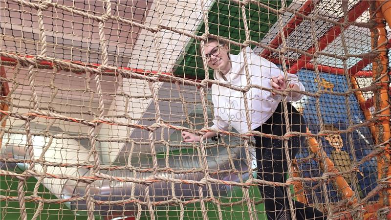 Person in weißem Hemd klettert in einem Netz aus Seilen in einer Indoor-Spielanlage.
