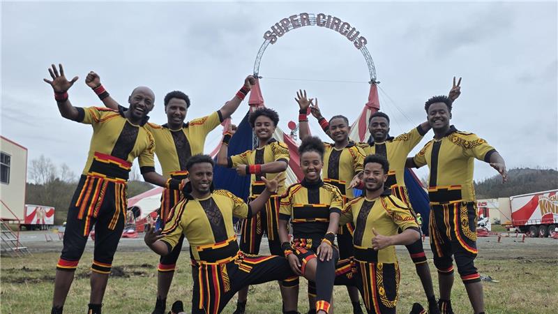 Gruppe von neun Personen in gelb-schwarzen Kostümen posiert vor einem Zirkuszelt mit Schriftzug 'SUPER CIRCUS'.