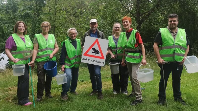 Sieben Personen mit grünen Warnwesten stehen auf einer Wiese vor Bäumen und halten Eimer und ein Schild mit der Aufschrift 'Krötenwanderung' und einem Dreieck-Warnzeichen mit einer Kröte.