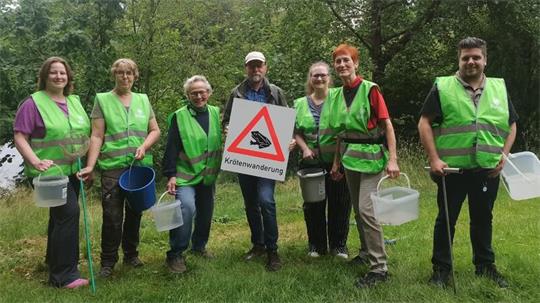 Sieben Personen mit grünen Warnwesten stehen auf einer Wiese vor Bäumen und halten Eimer und ein Schild mit der Aufschrift 'Krötenwanderung' und einem Dreieck-Warnzeichen mit einer Kröte.