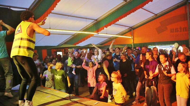 Menschen im Partyzelt schauen auf die Bühne auf der ein Künstler in Warnjacke gekleidet steht.