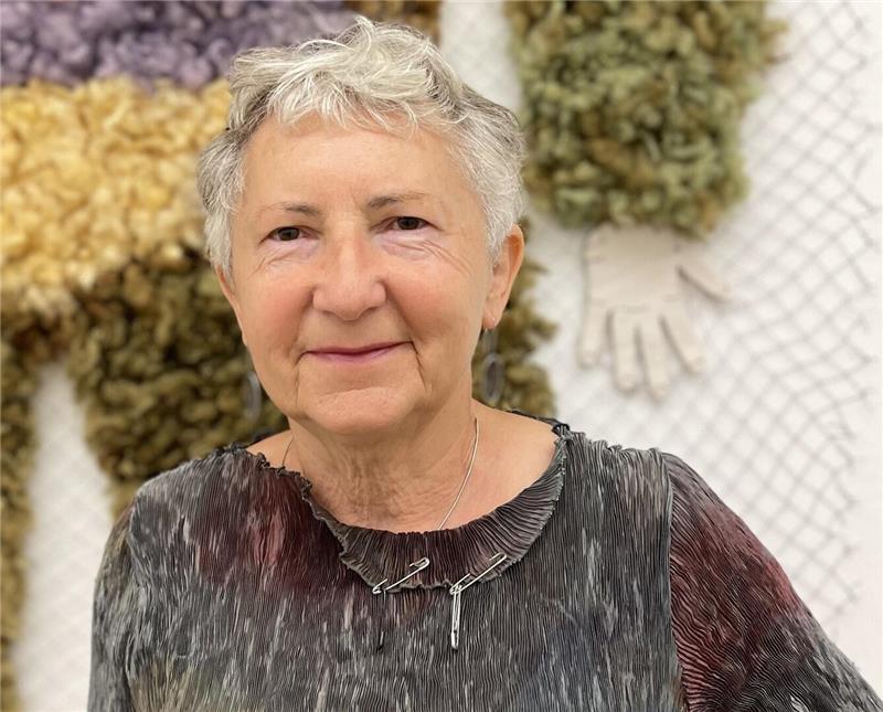 Eine Frau mit kurzen Haaren und sympathisch lächelndem Gesicht blickt den Betrachter an, hinter ihr eine Kunstarbeit aus Wolle.