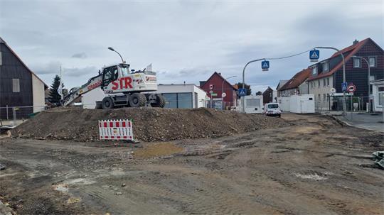 Baustelle mit Erdhügel und Bagger in einer Wohnstraße mit Häusern und Autos im Hintergrund