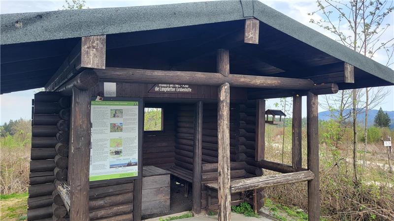 Holzunterstand mit Informationsschild in einer ländlichen Umgebung und bewaldeten Hügeln im Hintergrund