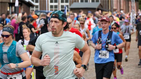 Läufer in Sportkleidung mit Startnummern bei einem Straßenlauf vor einem Gebäude mit Holzfassade